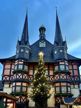 Yarı keresteli Wernigerode Town Hall, Wernigerode, Almanya, mavi gökyüzüne karşı iki noktalı kuleye sahiptir. Önünde yıldız tepesi olan uzun, aydınlık bir Noel ağacı duruyor.. 