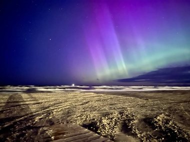 Görüntü, Letonya 'nın Liepaja kentindeki Baltık Denizi üzerinde kuzey ışıklarını yakalıyor. Gökyüzü mor ve yeşil auroraların canlı renkleriyle dolu. Ufuk boyunca uzanıyor.. 