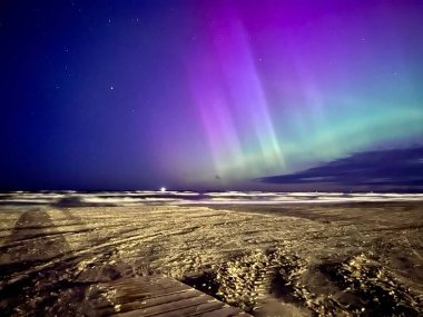 Görüntü, Letonya 'nın Liepaja kentindeki Baltık Denizi üzerinde kuzey ışıklarını yakalıyor. Gökyüzü mor ve yeşil auroraların canlı renkleriyle dolu. Ufuk boyunca uzanıyor.. 