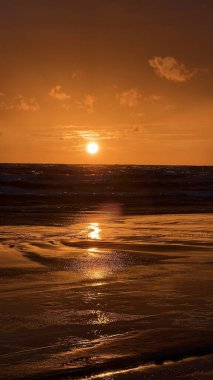 Güneş ufka yaklaştıkça deniz üzerinde gün batımı çarpıcı bir manzara yaratır. Gökyüzü ve su boyunca turuncu bir parıltı yayar. Küçük bulutlar üzerine dağılmış, sıcak renkleri yansıtıyor. Deniz yumuşak dalgalar sergiliyor