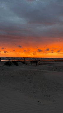 Letonya 'nın Liepaja kentindeki Baltık Denizi üzerinde gün batımı gökyüzünde canlı turuncu ve mor bir renk bırakıyor. Kara bulutlar ufku kısmen kaplayarak, manzaraya kontrastlık katıyor. Sakin deniz günbatımının ılık renklerini yansıtır.. 