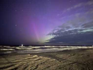 Kuzey ışıkları, Letonya 'nın Liepaja kentindeki Baltık Denizi üzerinde gece gökyüzünü aydınlatır. Ekranda mor ve yeşil renkli canlı renkler ve yıldızların arka planı yer alıyor.. 