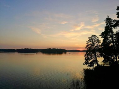 Günbatımı sakin bir göl üzerinde portakal, pembeler ve mavilerden oluşan eğimli bir gökyüzü oluşturur. Siluetli ağaçlar ön planı sağda çerçevelerken, uzaktan küçük bir tekne görülebiliyor ve ölçek ve sükunet hissi ekleniyor.. 