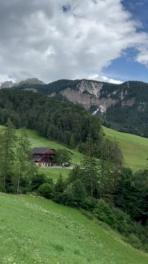 İtalya 'nın Dolomites kentindeki St. Magdalena köyünde yeşil tepeler ve geleneksel dağlık evler sergileniyor. Çan kulesine sahip seçkin bir kilise sahnenin merkezinde yer alıyor..