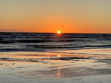 Letonya, Liepaja 'da Baltık Denizi üzerinde gün batımı. Güneş ufukta görünür, sıcak turuncu ve sarı bir parıltı yayar gökyüzünde. Yumuşak dalgalar kumlu kıyı şeridine doğru dalgalanır, ıslak kumlar canlı renkleri yansıtır..