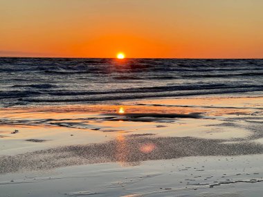 Sunset over the Baltic Sea in Liepaja, Latvia. The sun sits low on the horizon, casting a warm orange and yellow glow across the sky. Gentle waves ripple towards the sandy shoreline, where wet sand reflects the vibrant colors.