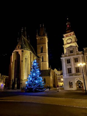 Çek Cumhuriyeti 'nin Hradec Kralove kentindeki gece sahnesinde karanlık bir gökyüzüne karşı aydınlatılmış tarihi bir kilise ve saat kulesi yer alıyor. Mavi ışıklarla süslenmiş büyük bir Noel ağacı şenlikli atmosferi güçlendirir..