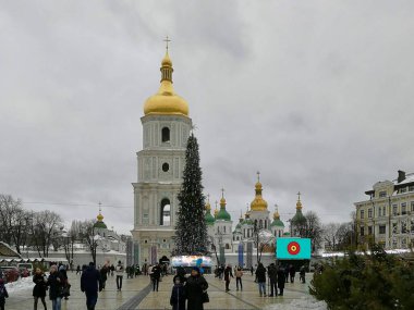 Ukrayna 'nın Kyiv kentindeki Aziz Sophia Katedrali' nin çan kulesi, süslü yapısının üzerinde belirgin bir altın kubbe barındırır. Görüntü, ön planda çok sayıda insanla çevrili büyük süslü bir Noel ağacının olduğu bir kış sahnesini çekiyor..