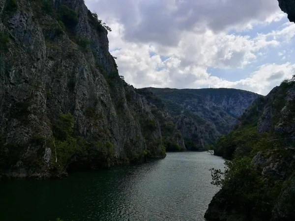 Makedonya 'nın kuzeyindeki Matka Kanyonu' ndaki dar, yeşil bir nehrin her iki yakasında da sarp kayalıklar yükseliyor. Uçurumlar karanlık, pürüzlü taşlarla çelişen bitki örtüsüyle kaplıdır. Parçalı bulutlu bir gökyüzü yayılmış güneş ışığına izin verir.