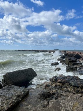 Dalgalar Liepaja, Letonya 'nın Baltık Denizi kıyısındaki kayalık bir kıyı şeridine çarpıyor. Sahnede koyu renkli kayalar ve parçalı bulutlu bir gökyüzünün altında çalkalanan bir deniz yer alıyor. Ufuk, suya uzanan taştan bir dalgakıran çizgisi gösteriyor.
