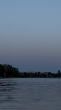 Yumuşak dalgalı sakin göl berrak mavi gökyüzünü yansıtıyor. Uzun otlar kıyı şeridi boyunca uzanır, sahneyi çerçeveler. Gölün karşısında, yoğun yeşil ağaçlar karşı kıyıyı kaplıyor.