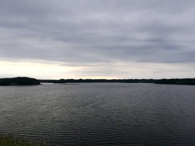 Litvanya, Zarasai 'de sakin bir göl, bulutlu bir gökyüzünün altında hafif bir ışık kırılması ile. Sakin su, yukarıdaki bulutların gri tonlarını yansıtıyor. Ufukta görünen yoğun ağaçlarla çevrili kıyı, açık sulara kontrastlık ekliyor.