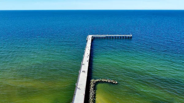 Palanga, Litvanya 'nın Baltık Denizi' ni gösteren hava manzarası. Koyu mavi sulara doğru uzanan uzun bir ahşap iskele L şeklinde bir şekil yaratır. Kıyı şeridi sakin dalgalar içerirken, iskele yürüyen insanlarla dolu..