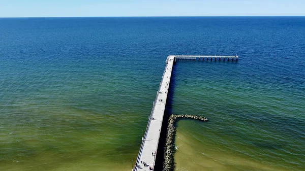 Palanga, Litvanya 'nın Baltık Denizi' ni gösteren hava manzarası. Koyu mavi sulara doğru uzanan uzun bir ahşap iskele L şeklinde bir şekil yaratır. Kıyı şeridi sakin dalgalar içerirken, iskele yürüyen insanlarla dolu..