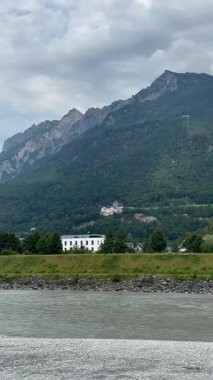 Vaduz 'daki şatonun manzarası. Lihtenştayn Prensi 'nin evi. Resimli bir manzara, yemyeşil bir yamaçta yuva yapmış bir kaleyi gözler önüne seriyor. Ön planda, kayalık kıyılar arasında küçük bir nehir akar ve bir kişi kanoda kürek çeker..