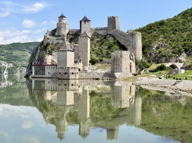 Sırbistan 'daki Golubac Kalesi Tuna Nehri' nin yanındaki kayalık bir tepeye tünemiş durumda. Ortaçağ yapısı, nehrin sakin sularına güzel yansıyan, konik çatıları ve taş duvarları olan birden fazla kuleyi sergiliyor. Gökyüzü açık