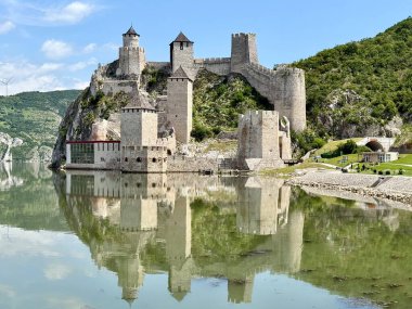 Sırbistan 'daki Golubac Kalesi Tuna Nehri' nin yanındaki kayalık bir tepeye tünemiş durumda. Ortaçağ yapısı, nehrin sakin sularına güzel yansıyan, konik çatıları ve taş duvarları olan birden fazla kuleyi sergiliyor. Gökyüzü açık