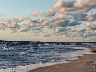 Nazik dalgalar, Litvanya 'nın Baltık Denizi yakınlarındaki Palanga' nın kumlu kıyılarına vuruyor. Batan güneşle aydınlanan kabarık bulutlar suyun üzerine yumuşak bir ışık saçıyor. Deniz, sabahtan akşama geçişi yansıtan hafif dalgalı görünüyor..