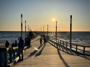 Görüntü Palanga, Litvanya 'da gün batımında Baltık Denizi' ne uzanan ahşap bir iskeleyi gösteriyor. Gökyüzü açık ve güneş ufukta görünüyor. İskelede yürüyen insanların uzun gölgelerini oluşturuyor. Işık, sahne boyunca sıcak, altın bir parıltı yaratır.