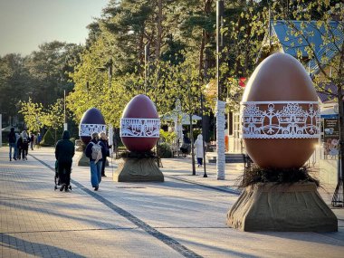 Süslü dev Paskalya yumurtaları Litvanya 'nın Palanga kentindeki asfalt bir patika boyunca sergilenir. Her yumurta karmaşık beyaz dantel desenlerle bezenmiş, binalar görünür durumda, Paskalya kutlamalarındaki hareketli sokak manzarasını güçlendiriyor..