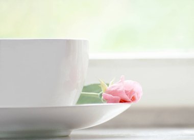 Pencere kenarında pembe gül goncası olan beyaz bardak.