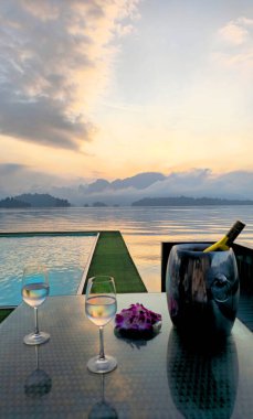 Sonsuzluk havuzuna bakan iki bardak ve şişe masa ve gün batımında Cheow Lan Gölü, Khao Sok Ulusal Parkı, Tayland, Güneydoğu Asya.     