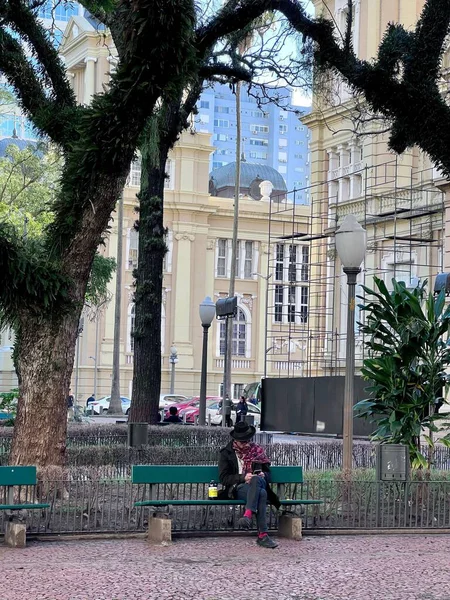 Bir adam parktaki bir bankta oturuyor, faaliyetlerine dalmış. Sahne, arka planda yemyeşil ağaçlar ve mimari binalarla çevrili, sakin ve huzurlu bir günü resmediyor..