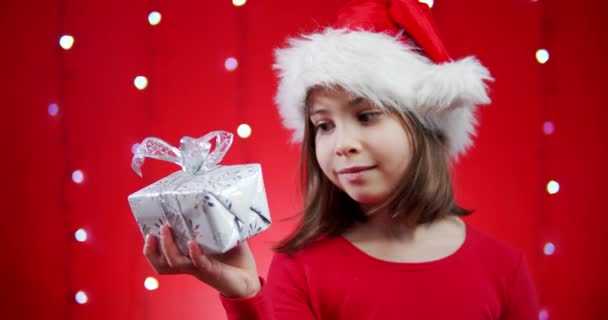 Słodkie dziewczyna w Santa kapelusz posiada prezent na czerwonym tle Bożego Narodzenia — Wideo stockowe