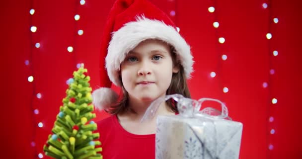 Мила дівчина в капелюсі Санта тримає подарунок на червоному різдвяному фоні — стокове відео