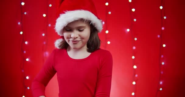 Menina bonito em um chapéu de santa abre um presente em um fundo de Natal vermelho — Vídeo de Stock