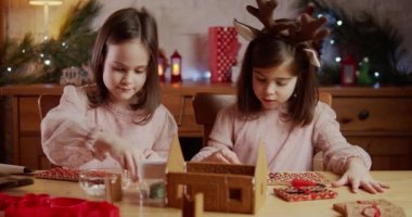İki tatlı kız kardeş bir Noel kurabiyesi evi yapıp dekore ediyorlar.