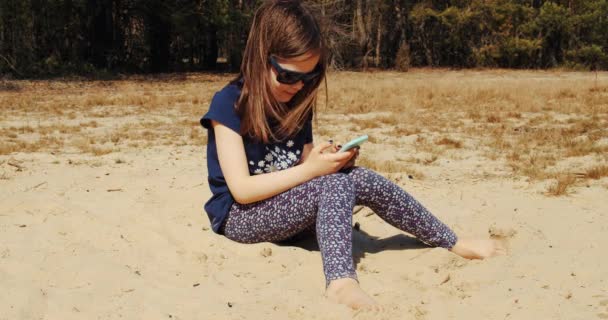 Милая маленькая девочка держит телефон в руке и сидит в летнем лесу — стоковое видео