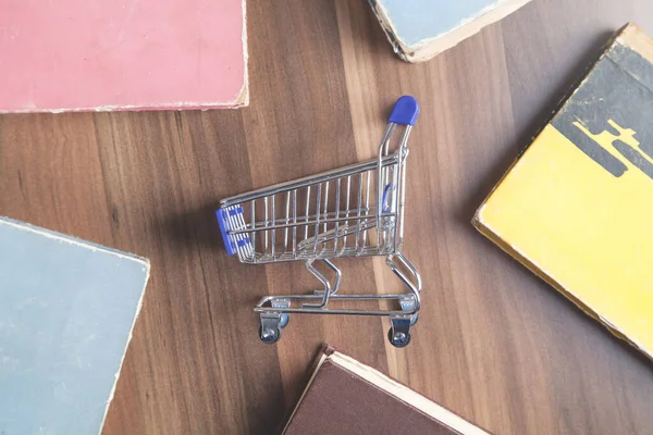 Shopping cart and books on wooden background.