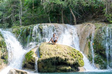 A man sits peacefully on a mossy rock surrounded by a tropical waterfall and lush forest. A serene travel and adventure photo symbolizing freedom, exploration, and connection with nature.