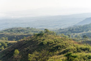 Puslu bir sabah gökyüzünün altında yuvarlanan yeşil ve kahverengi tepelerden oluşan engin bir manzarayı gösteren güzel bir fotoğraf. Yumuşak ışık, sakin ve geniş kırsal manzarayı aydınlatır..