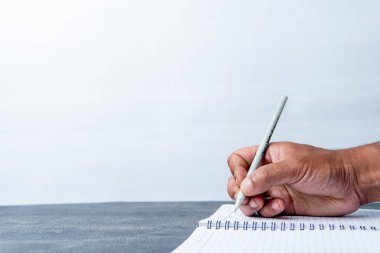 Gri bir masadaki sarmal bir defterde kalemle yazan bir adamın yakın plan fotoğrafı. Temiz beyaz arka plan yeterli kopya alanı sağlar. Çalışmayı, çalışmayı ya da planlamayı temsil eder.