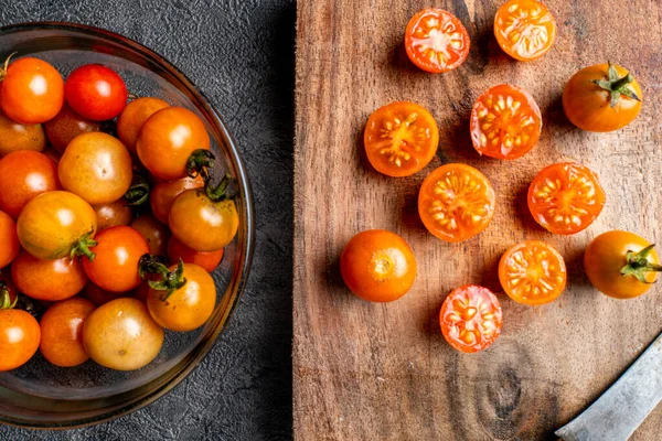Taze vişneli domateslerin üst görünümü. Bir cam kase dolusu domates koyu bir arka planda, domateslerin yanında, kırsal bir ahşap kesim tahtasının üzerinde. Yemek pişirmek ya da sağlıklı bir yemek tarifi.