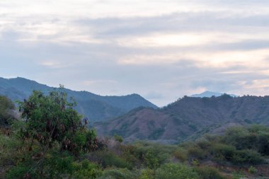 Geniş bir dağ vadisi manzarası. Vadi yeşil çalılarla dolu. Güneş parlak, puslu bir atmosfer yaratıyor..