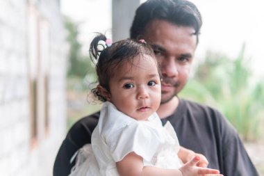 Sevgi dolu bir Asyalı baba tapılası bebeğini kucağına alır. Meraklı kız, beyaz elbiseli, doğrudan kameraya bakıyor. Sıcak, samimi bir aile anı..