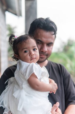 Sevgi dolu bir Asyalı baba tapılası bebeğini kucağına alır. Meraklı kız, beyaz elbiseli, doğrudan kameraya bakıyor. Sıcak, samimi bir aile anı..