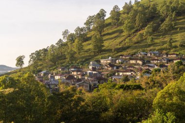 Ağaçlarla çevrili güneşli, sarp, yeşil yamaçlara yerleşmiş geleneksel evleri gösteren yüksek bir manzara..