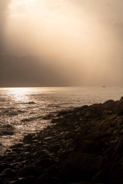 Karanlık, kasvetli bir kayalık kıyı şeridinde gizemli güneş ışınları bulutları delip geçiyor. Uçsuz bucaksız okyanus ufkunda bir teknenin silueti..