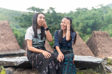 Bena Village 'da geleneksel sazdan çatı evleri ve yeşil tepelerle sohbet eden iki gülümseyen kadın..