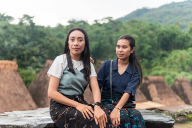 Bena Village 'da geleneksel sazdan yapılmış evlerle birlikte etek dokuma elbiseler giyen iki gülümseyen Asyalı kadın dışarıda oturuyor..