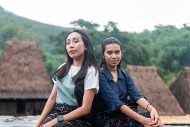 Bena Village 'da geleneksel sazdan yapılmış evlerle birlikte etek dokuma elbiseler giyen iki gülümseyen Asyalı kadın dışarıda oturuyor..