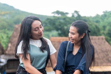 Bena Village 'da geleneksel sazdan çatı evleri ve yeşil tepelerle sohbet eden iki gülümseyen kadın..