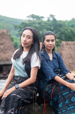 Bena Village 'da geleneksel sazdan yapılmış evlerle birlikte etek dokuma elbiseler giyen iki gülümseyen Asyalı kadın dışarıda oturuyor..