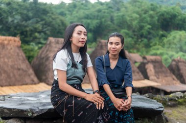 Bena Village 'da geleneksel sazdan yapılmış evlerle birlikte etek dokuma elbiseler giyen iki gülümseyen Asyalı kadın dışarıda oturuyor..