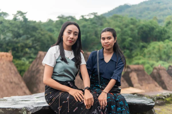 Bena Village 'da geleneksel sazdan yapılmış evlerle birlikte etek dokuma elbiseler giyen iki gülümseyen Asyalı kadın dışarıda oturuyor..