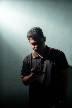 Akıllı telefonuna odaklanmış genç bir adamın ön görüntüsü, arka planda koyu dokulu bir ışıkla aydınlatılmış..